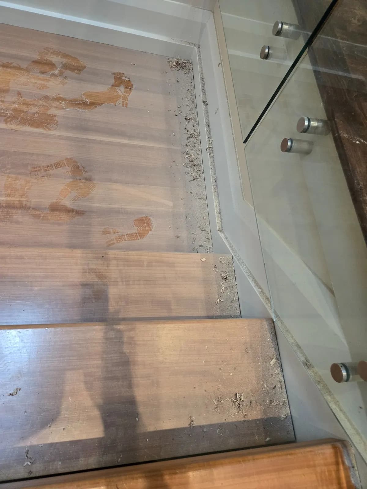 Loose wood shavings and fine dust gathered along the edges of wooden stair treads, beside a glass railing, before cleaning